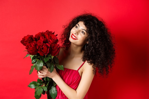 Foto feliz hermosa mujer vestida, sosteniendo flores y sonriendo romántico, mirando a un lado en el logo, de pie contra el fondo rojo.