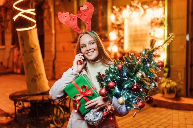 Feliz y hermosa niña caucásica sosteniendo un árbol de Navidad y hablando por teléfono