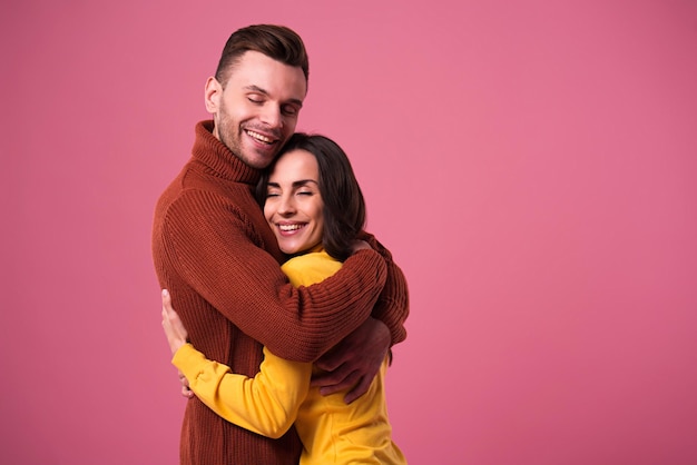 Feliz hermosa pareja amorosa vestida con suéteres abrazándose aislado sobre fondo rosa