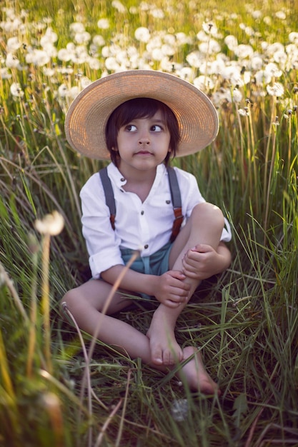 Feliz, un hermoso niño con sombrero se para en un campo con dientes de león blancos al atardecer en verano