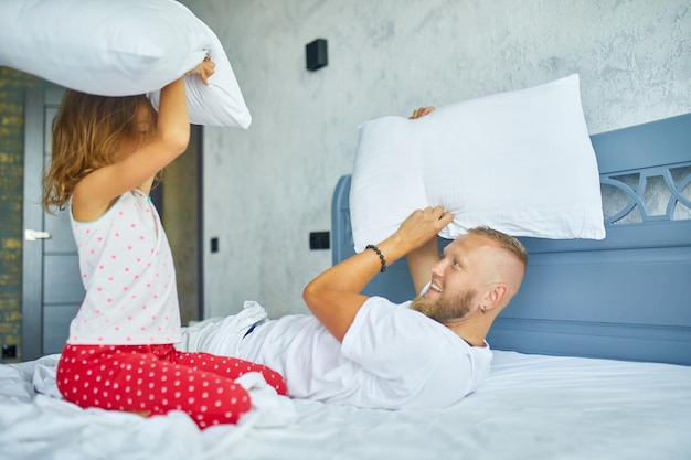 Feliz hija y padre jugando a la pelea de almohadas en la cama por la mañana en casa, peleando con almohadas, divertirse jugar juntos en casa el fin de semana familiar