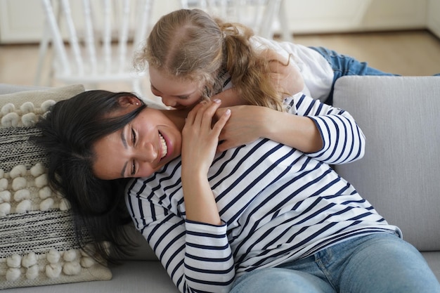 Feliz hijita haciéndole cosquillas a mamá o niñera sentada en el sofá divirtiéndose juntos