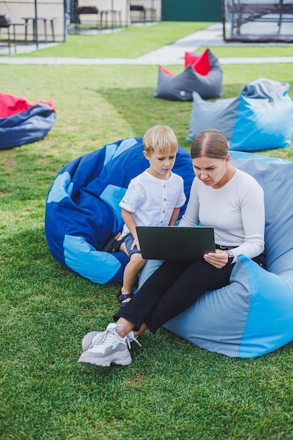 Feliz hijo y madre sentados en la naturaleza y mirando la computadora portátil Trabajo remoto de una joven madre Vacaciones familiares