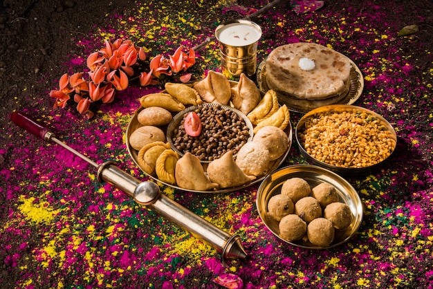 Foto feliz holi cartão projetado mostrando comida tradicional indiana doce e salgada, flores e cores em pó dispostas sobre fundo branco ou argila. foco seletivo