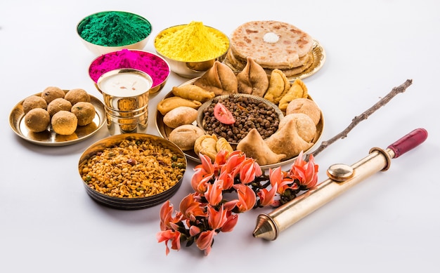 Feliz Holi cartão projetado mostrando comida tradicional indiana doce e salgada, flores e cores em pó dispostas sobre fundo branco ou argila. Foco seletivo