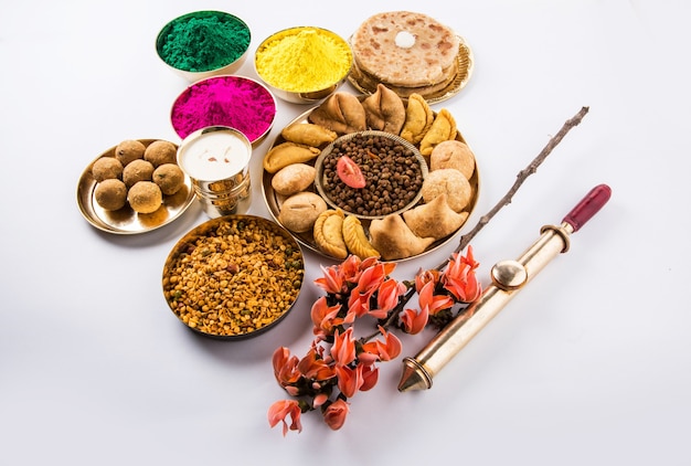 Feliz Holi cartão projetado mostrando comida tradicional indiana doce e salgada, flores e cores em pó dispostas sobre fundo branco ou argila. Foco seletivo
