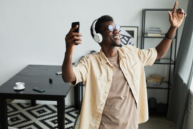 Feliz hombre africano en auriculares inalámbricos con su teléfono móvil para escuchar música y bailar en el ...