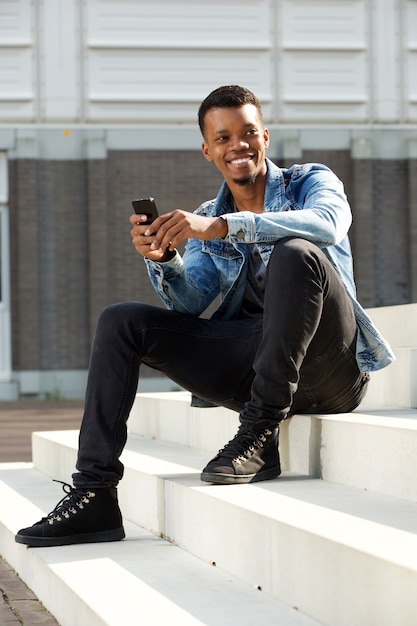 Feliz hombre africano sentado en pasos afuera sosteniendo teléfono inteligente