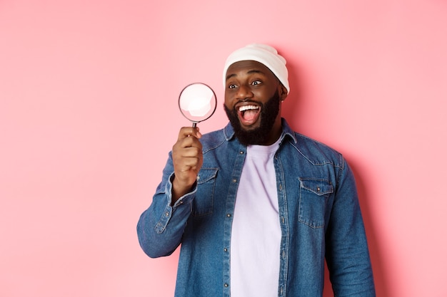 Feliz hombre afroamericano mirando a través de una lupa, sonriendo asombrado, de pie contra el fondo de color rosa. Copia espacio