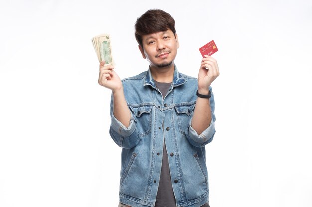 Foto feliz hombre asiático en la cara sonriente sosteniendo una tarjeta de crédito y un billete de dólar, mira a la cámara, retrato de luz de estudio aislado sobre fondo blanco, concepto de tarjeta de crédito