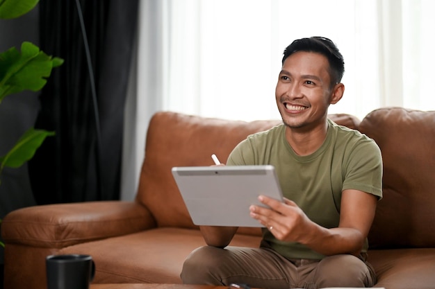 Feliz hombre asiático milenario usando su tableta digital mientras ve la televisión en la sala de estar