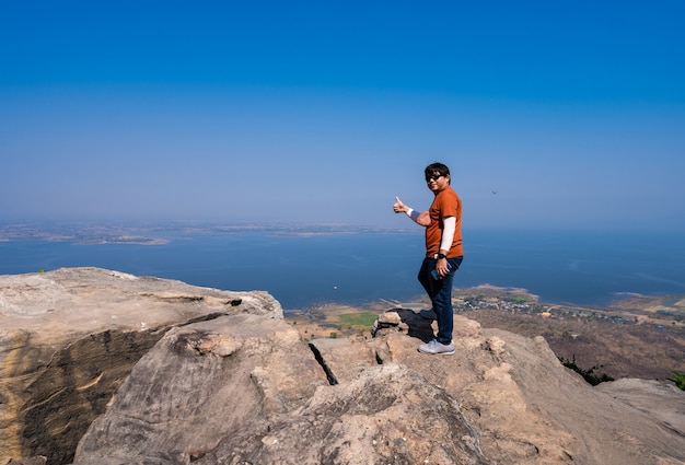 Feliz hombre asiático de pie con sonrisa feliz y pulgar hacia arriba en el mirador superior en el pico de la montaña de roca con vista a la presa del lago brumoso y cielo azul en el mirador de Pha Muen, Khon Kaen, Tailandia, satisfacer el concepto de viaje