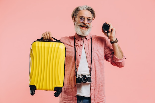 Feliz hombre barbudo con estilo en gafas de sol con cámara sosteniendo una maleta y un altavoz portátil