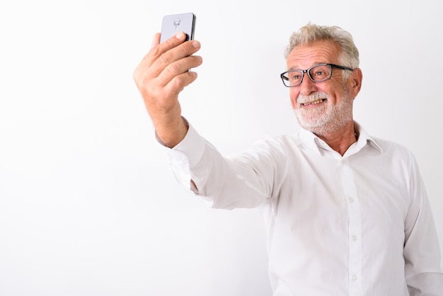 Feliz hombre barbudo senior sonriendo mientras toma una foto selfie con teléfono móvil mientras usa anteojos en blanco