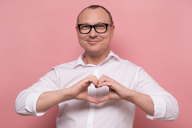 Feliz hombre caucásico maduro con gafas que muestra el símbolo del corazón en el fondo rosa