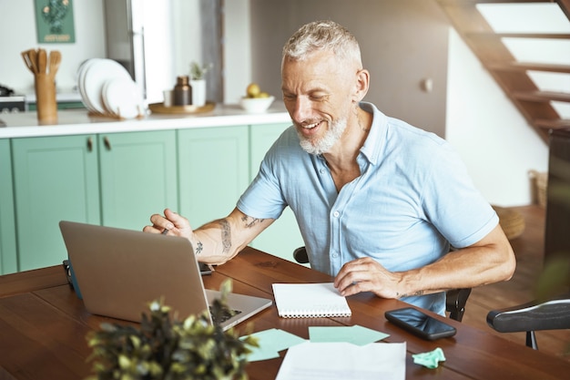 Feliz hombre caucásico de mediana edad que usa la computadora portátil mientras está sentado en la mesa y trabaja en línea desde casa