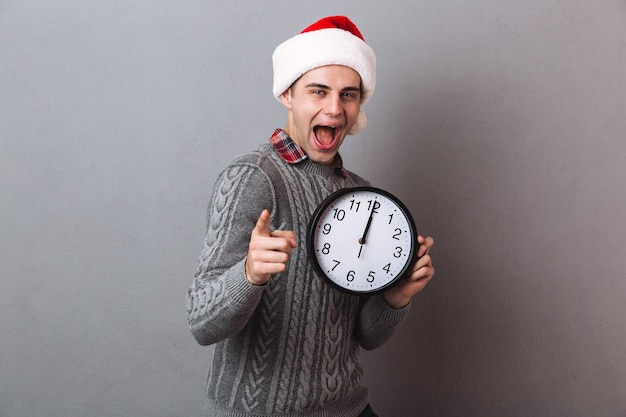 Feliz hombre gritando en suéter y sombrero de navidad sosteniendo reloj y apuntando