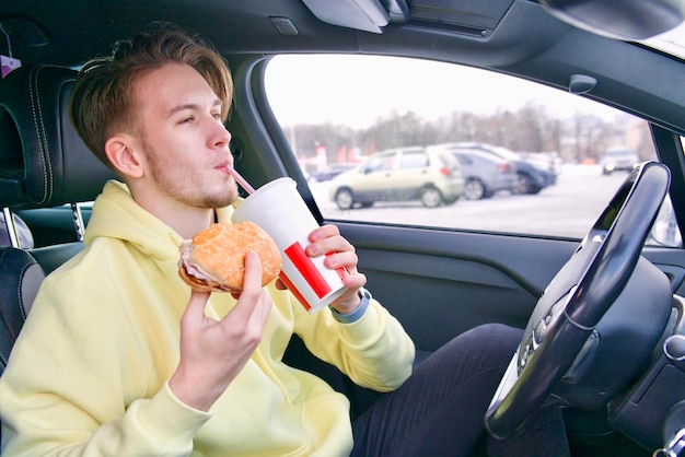 feliz hombre hambriento joven conductor sentado en el coche comiendo hamburguesas bebiendo refrescos comida rápida