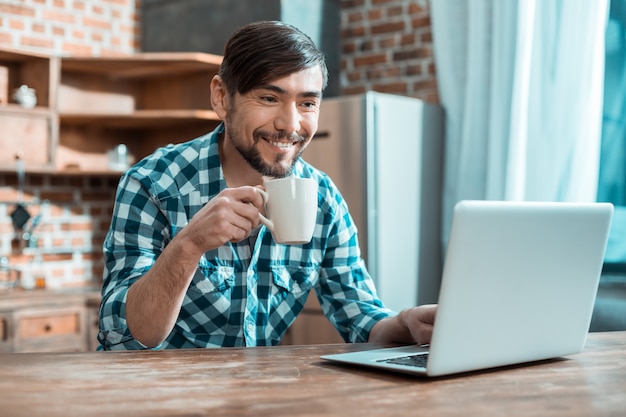 Feliz hombre inteligente positivo sentado frente a la computadora portátil y tomando té mientras trabaja desde casa