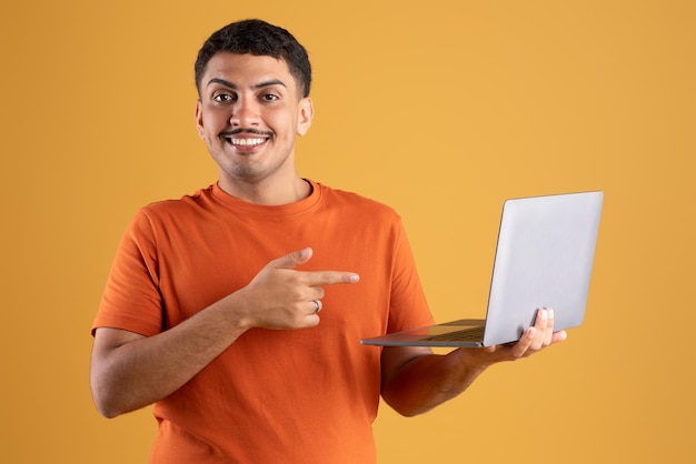 Feliz hombre latino apuntando a la pantalla del portátil y sonriendo de pie con la PC en el fondo amarillo del estudio