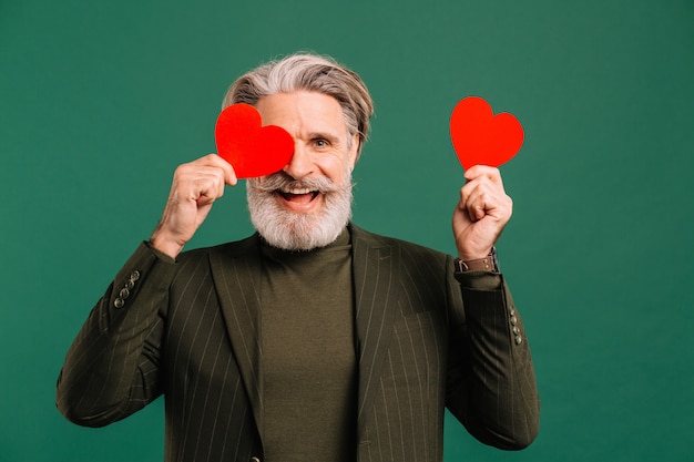 Feliz hombre maduro con barba y bigote en traje de color caqui con dos corazones rojos cerca de los ojos que saludan a San Valentín isoleted fondo verde.