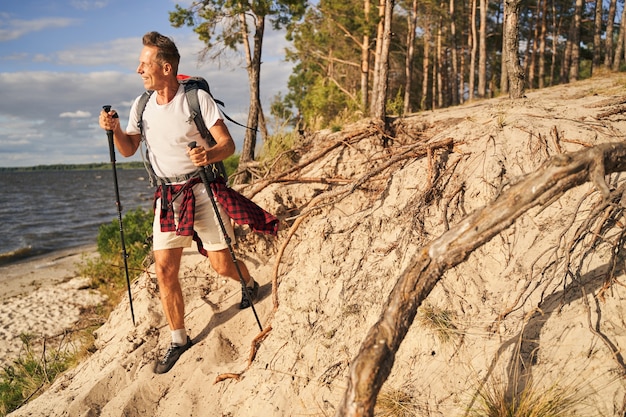 Feliz hombre maduro deportivo va nordic walking con mochila en el bosque cerca de la orilla del mar