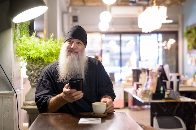 Feliz hombre maduro hipster barbudo pensando mientras usa el teléfono en la cafetería.