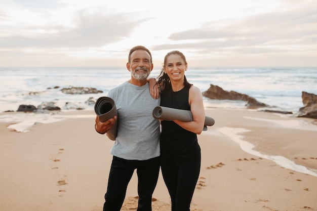 Feliz hombre y mujer caucásicos maduros en ropa deportiva con colchonetas listas para hacer ejercicio en la mañana en el mar