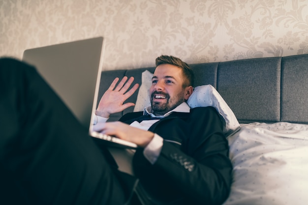 Feliz hombre de negocios acostado en la cama en la habitación del hotel en traje y usando la computadora portátil para videollamadas.