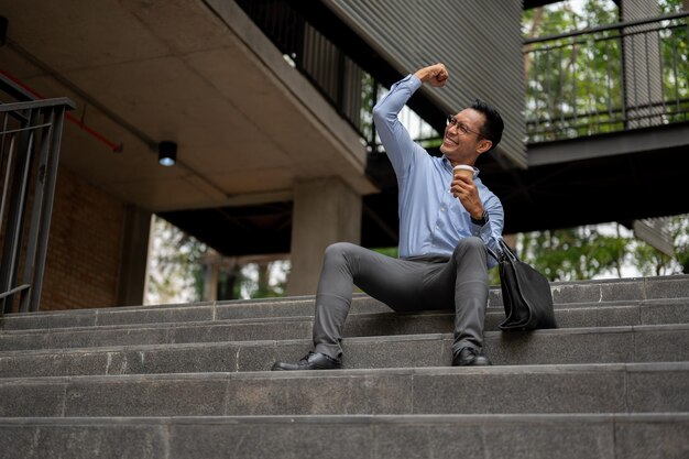 Foto un feliz hombre de negocios asiático se sienta en los escalones con una taza de café y aplaude con el puño levantado