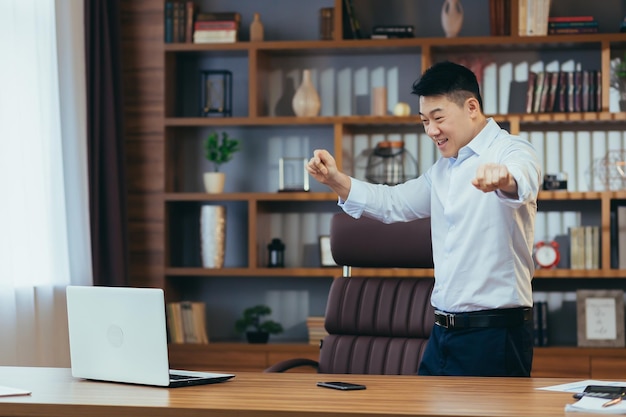 Feliz hombre de negocios bailando y celebrando la victoria sentado en una mesa en una oficina clásica asiática trabajando en la computadora