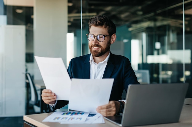 Feliz hombre de negocios leyendo documentos e informes comerciales regocijándose en los asuntos de la empresa sentados en una oficina moderna