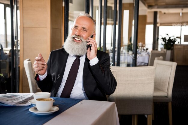 Feliz hombre de negocios maduro hablando por teléfono en el restaurante mientras espera la reunión