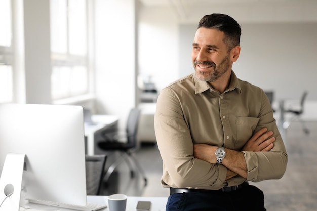 Feliz hombre de negocios de mediana edad de pie junto a su lugar de trabajo mirando a un lado el espacio de copia pensando en una nueva empresa o proyecto