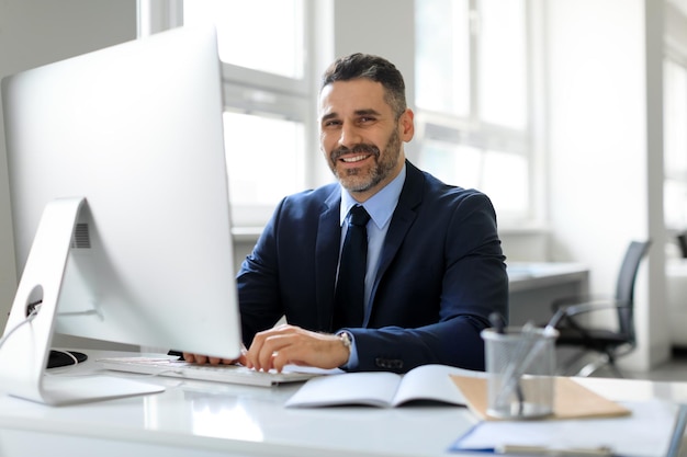 Feliz hombre de negocios de mediana edad en traje sentado en la oficina y sonriendo a la cámara trabajando con espacio libre de computadora moderna