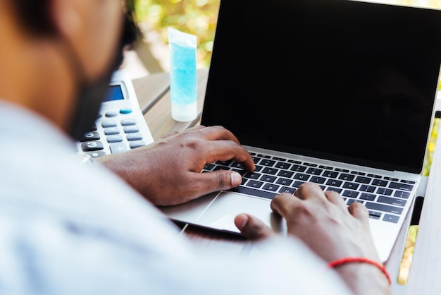 Un feliz hombre de negocios negro asiático o un trabajador de oficina usan máscara facial para protegerse sentados en el escritorio desde casa usando el teclado de una computadora portátil y conectando redes en línea en la cafetería