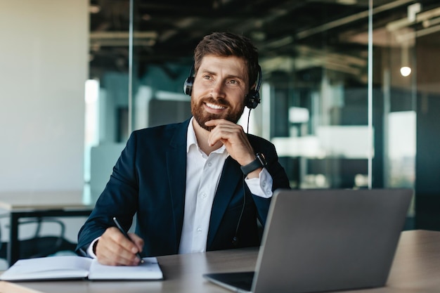 Feliz hombre de negocios pensativo en auriculares trabajando en una computadora portátil y escribiendo en un cuaderno sentado en el escritorio en el interior de la oficina