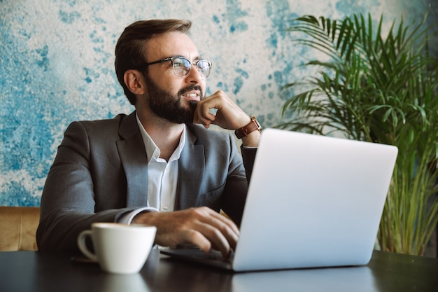 Feliz hombre de negocios trabajando en una computadora portátil mientras está sentado en la cafetería y tomando café en el interior