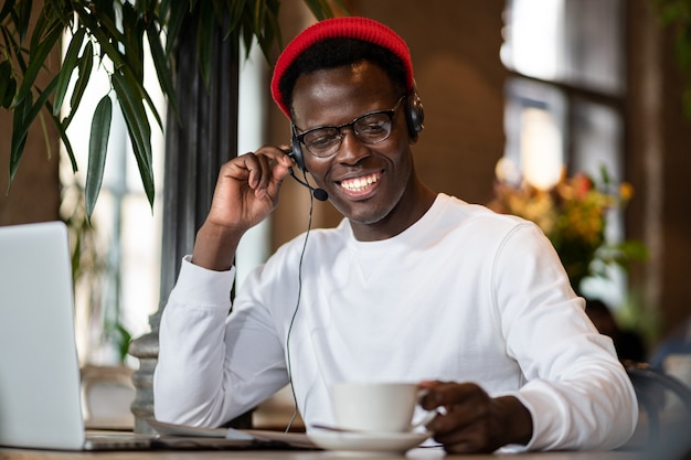 Feliz hombre negro milenario en auriculares viendo webinar en portátil, trabajo en línea de forma remota en café, sosteniendo una taza de café