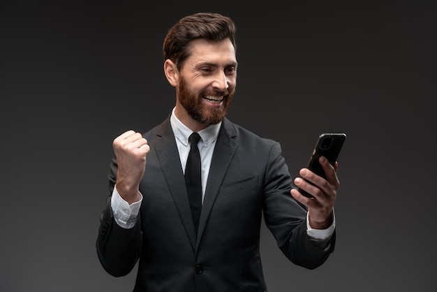 Feliz hombre satisfecho con barba sosteniendo un teléfono inteligente y sonriendo mientras hace un gesto de sí, celebrando la lotería en línea o la victoria del sorteo. Tiro de estudio interior aislado sobre fondo negro