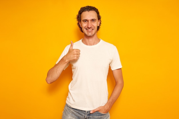 Feliz el hombre sonriente en ropa casual con camiseta blanca sobre fondo amarillo muestra Thumbs up likes