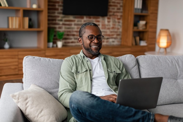 Feliz homem afro-americano adulto em óculos assistindo vídeo no estudo do laptop desfruta de descanso no tempo livre sozinho