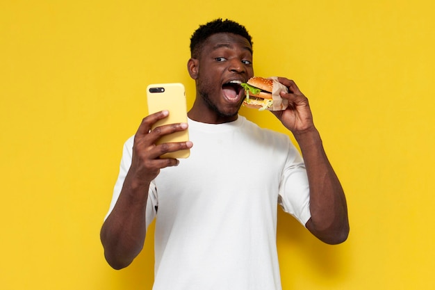 feliz homem afro-americano em camiseta branca, segurando um hambúrguer grande e usando o smartphone, o cara está comendo