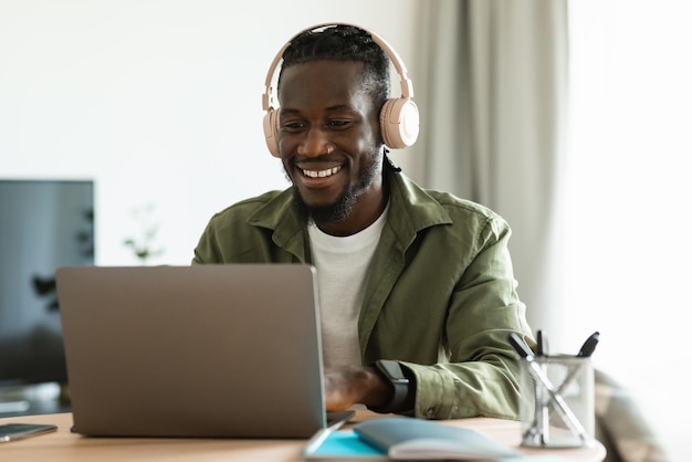 Feliz homem afro-americano no fone de ouvido trabalhando no laptop sentado no escritório em casa e consultando clientes on-line