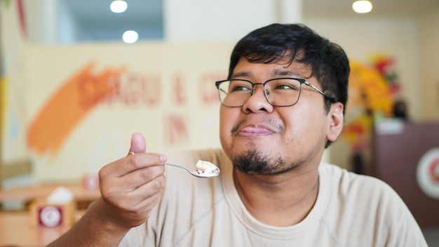 Foto feliz homem asiático com óculos mordendo o lábio e comendo um delicioso sorvete com apetite desfrutando do sabor natural dentro do restaurante