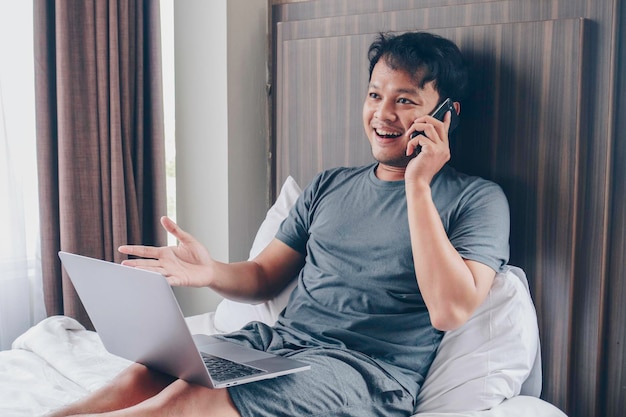 Feliz homem asiático está trabalhando com seu laptop com videochamada em uma cama Conceito de estilo de vida bem-sucedido freelancer