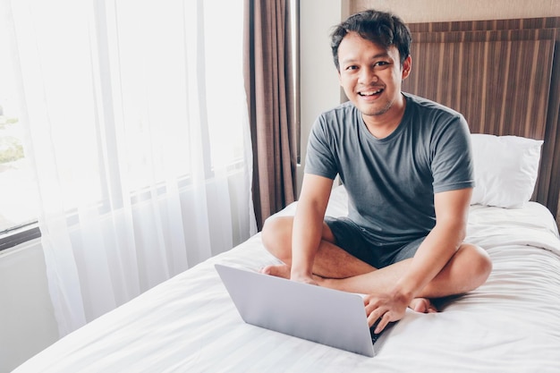 Feliz homem asiático está trabalhando com seu laptop em sua cama Conceito de estilo de vida bem-sucedido freelancer