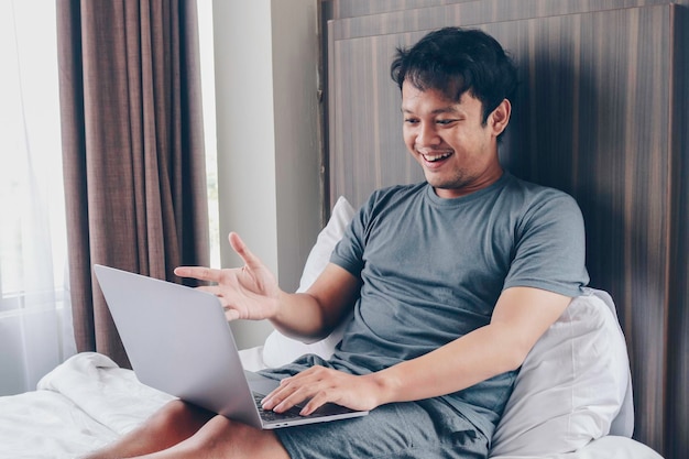 Feliz homem asiático está trabalhando com seu laptop em sua cama Conceito de estilo de vida bem-sucedido freelancer