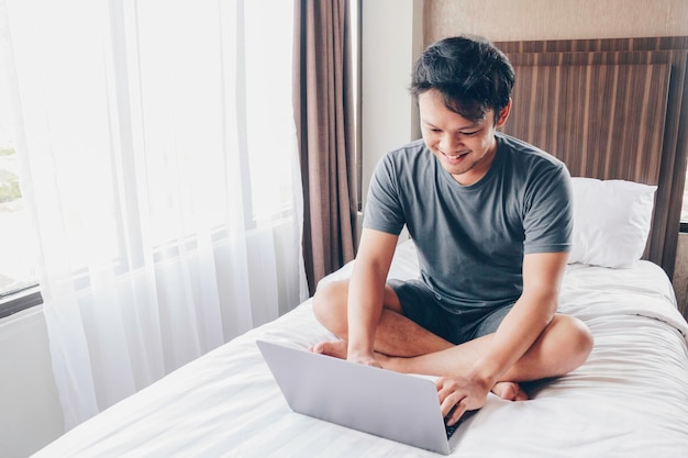 Feliz homem asiático está trabalhando com seu laptop em sua cama conceito de estilo de vida bem-sucedido freelancer