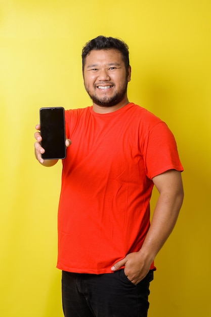 Feliz homem asiático gordo de camisa vermelha sorrindo enquanto segura smartphone isolado em fundo amarelo